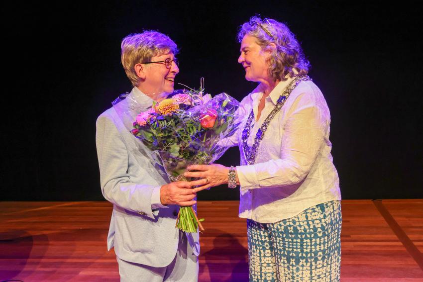 Jan Goverde ontvangt de onderscheiding en bloemen van locoburgemeester Mieke Verhees.